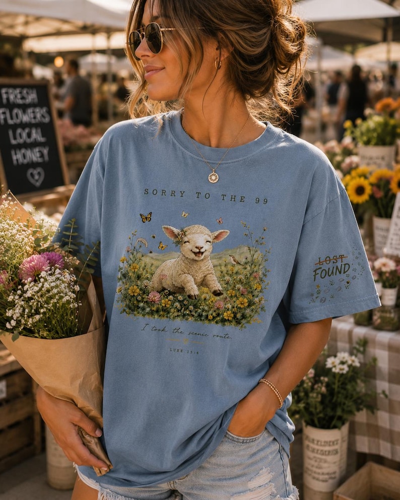 May include: A light blue t-shirt featuring a graphic of a smiling lamb sitting in a field of colorful wildflowers and butterflies. The text reads Sorry to the 99, Lost Found, I took the scenic route, and Luke 15:4. The shirt has a crew neckline.