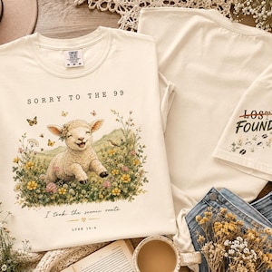 May include: A cream-colored t-shirt featuring a graphic of a smiling lamb in a field of flowers with butterflies. The text reads SORRY TO THE 99, I took the scenic route, and LUKE 15:4. The sleeve displays the text LOST FOUND with floral accents.