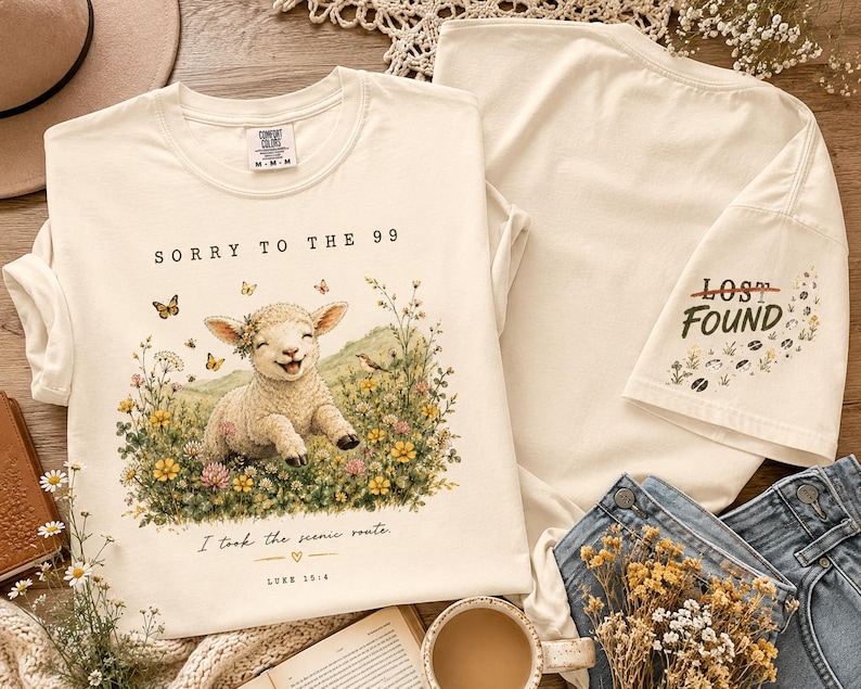 May include: A cream-colored t-shirt featuring a graphic of a smiling lamb in a field of flowers with butterflies. The text reads SORRY TO THE 99, I took the scenic route, and LUKE 15:4. The sleeve displays the text LOST FOUND with floral accents.