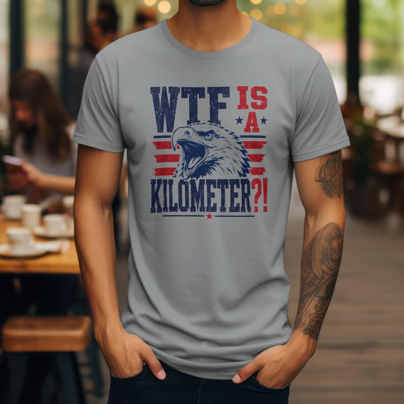 Pode incluir: Camiseta cinza com um gr&aacute;fico desgastado. O design apresenta a cabe&ccedil;a de uma &aacute;guia e o texto "WTF IS A KILOMETER?!" em azul marinho e vermelho. O design incorpora estrelas e listras, evocando um tema patri&oacute;tico.