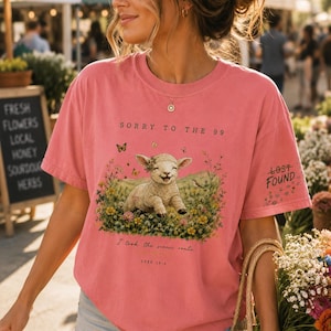May include: A coral pink short-sleeve t-shirt featuring a graphic of a lamb in a field of flowers with butterflies. The text reads SORRY TO THE 99, I took the scenic route, LUKE 15:4, and LOST FOUND on the sleeve. The shirt has a crew neckline.