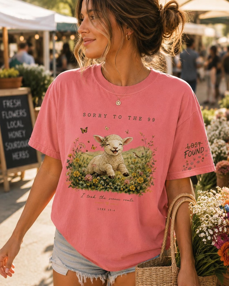 May include: A coral pink short-sleeve t-shirt featuring a graphic of a lamb in a field of flowers with butterflies. The text reads SORRY TO THE 99, I took the scenic route, LUKE 15:4, and LOST FOUND on the sleeve. The shirt has a crew neckline.
