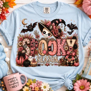 May include: A light blue t-shirt featuring a graphic print with the text "SPOOKY SEASON". The design includes a skeleton wearing a floral witch hat, bats, pumpkins, and flowers in shades of pink, black, and tan. The shirt has a Comfort Colors label.