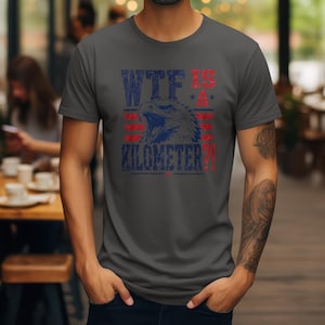 Pode incluir: T-shirt cinza escuro com um gr&aacute;fico desgastado com uma &aacute;guia azul e vermelha e o texto "WTF IS A KILOMETER?!" em azul e vermelho. A camisa tem mangas curtas e gola redonda.