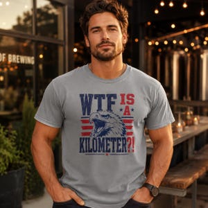 Pode incluir: Camiseta cinza com a frase "WTF IS *A* KILOMETER?!" em azul e vermelho, com um gr&aacute;fico de uma &aacute;guia. A camiseta &eacute; de gola redonda e parece ser feita de um material macio.