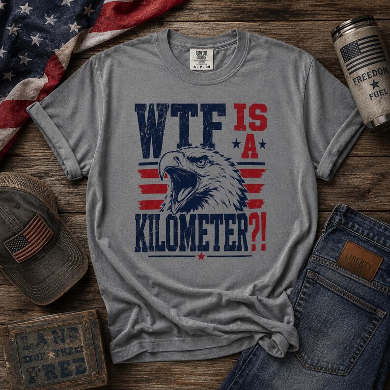 Pode incluir: Uma camiseta cinza com o texto "WTF IS A KILOMETER?!" e um gr&aacute;fico de &aacute;guia em vermelho e azul. A imagem inclui um bon&eacute; de beisebol com uma bandeira americana, uma lata com uma bandeira americana e jeans azuis.