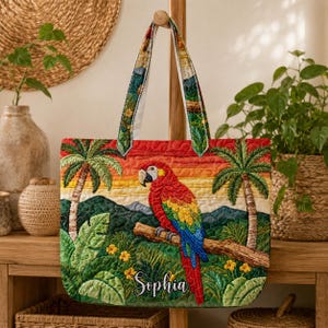 Könnte beinhalten: Eine gesteppte Tragetasche mit einem bunten Papagei auf einem Ast, vor einer tropischen Sonnenuntergangsszene mit Palmen und üppigem Laub. Die Tasche hat gemusterte Griffe und der Name "Sophia" ist auf der Vorderseite gestickt.