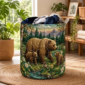 May include: A cylindrical fabric laundry hamper featuring a woven tapestry design of a large brown bear with two cubs in a forest landscape with mountains, trees, and flowers. The hamper has two white fabric handles and contains folded clothing.