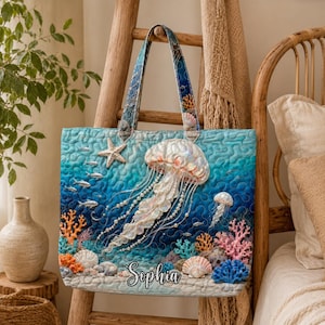 May include: A blue quilted tote bag featuring a jellyfish, starfish, and coral reef design. The bag has a personalized name, Sophia, and a blue and patterned handle. The bag is decorated with pearls and other sea creatures.