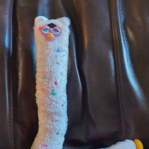 May include: A light pink plush toy in the shape of the letter "L". The toy has a Furby-like face with pink, blue, and yellow accents. It is decorated with colorful beads and small star-shaped sequins. The feet are yellow.