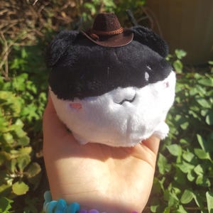 以下が含まれることがあります： 黒い上部と白い下部を持つ丸いぬいぐるみ。小さな刺繍のピンクの頬とシンプルなステッチの口が特徴で、細い編み込みバンドが付いた小さな茶色のフェルト製カウボーイハットをかぶっています。