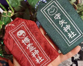 Puede incluir: Dos amuletos omamori japoneses. Uno es rojo con un símbolo Yin-Yang blanco y texto, el otro es verde con una ilustración de rana y texto. Ambos tienen bordes blancos y se sostienen en una mano.