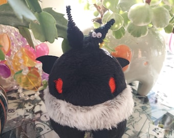 Puede incluir: Un peluche negro con forma de criatura con detalles rojos y una sección blanca y esponjosa. El juguete tiene pequeñas alas y antenas, sobre un fondo de plantas y un jarrón decorativo.