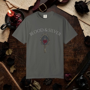 May include: Gray t-shirt with the text "BLOOD & SILVER" above a decorative mirror graphic. The mirror features a red rose and a sword. The shirt is made of a soft material.