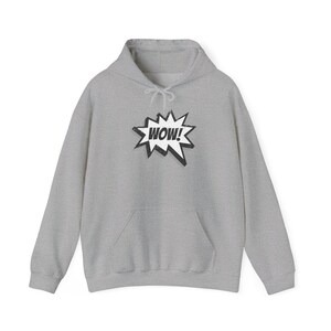 Puede incluir: Una sudadera con capucha gris claro con un bolsillo delantero y una capucha con cordón. El gráfico en la parte delantera presenta la palabra "WOW!" en un bocadillo de estilo cómic en blanco y negro. La sudadera con capucha está hecha de un material suave y cómodo.