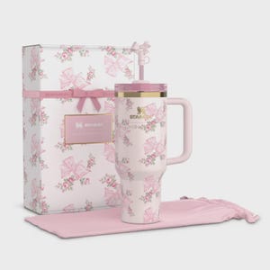 Puede incluir: Un vaso Stanley rosa y blanco con un estampado floral y lazos, una caja de regalo a juego y una funda de transporte rosa. El vaso tiene un asa y una pajita. La caja de regalo tiene una cinta rosa y una etiqueta dorada con "Stanley".