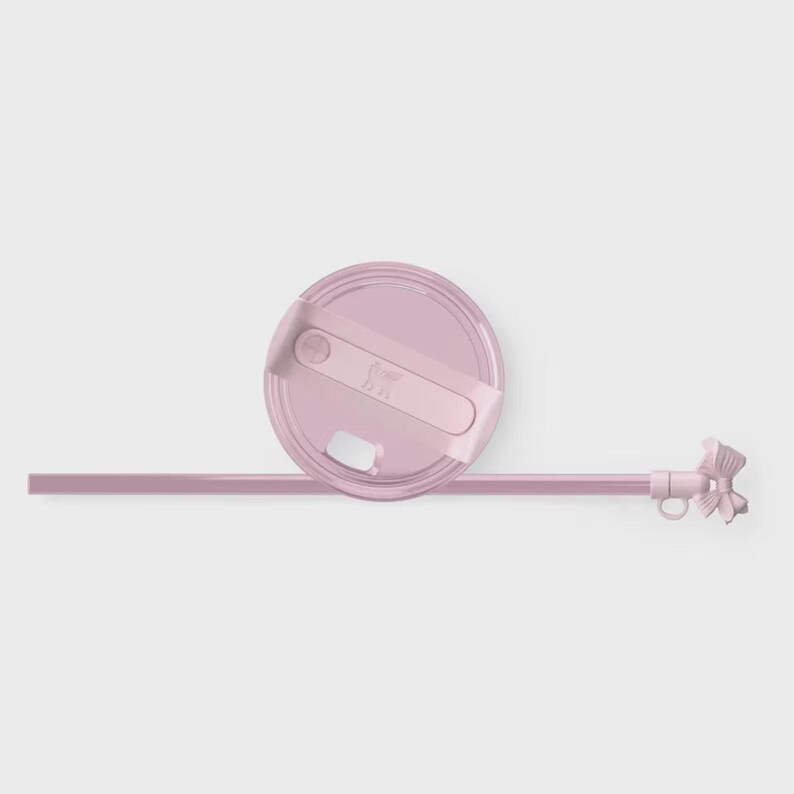 Puede incluir: Una tapa de vaso rosa claro con una pajita y un lazo a juego. La tapa tiene un cierre deslizante y un peque&ntilde;o logotipo. La pajita recta tiene un lazo decorativo en el extremo.