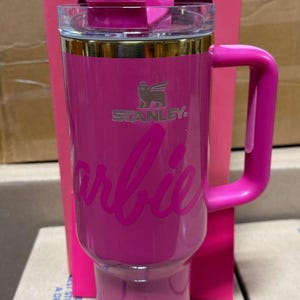 May include: A pink Stanley travel mug with the Barbie logo. The mug has a handle, a clear lid, and gold trim. The Stanley logo is also visible. The mug is in its original pink box.