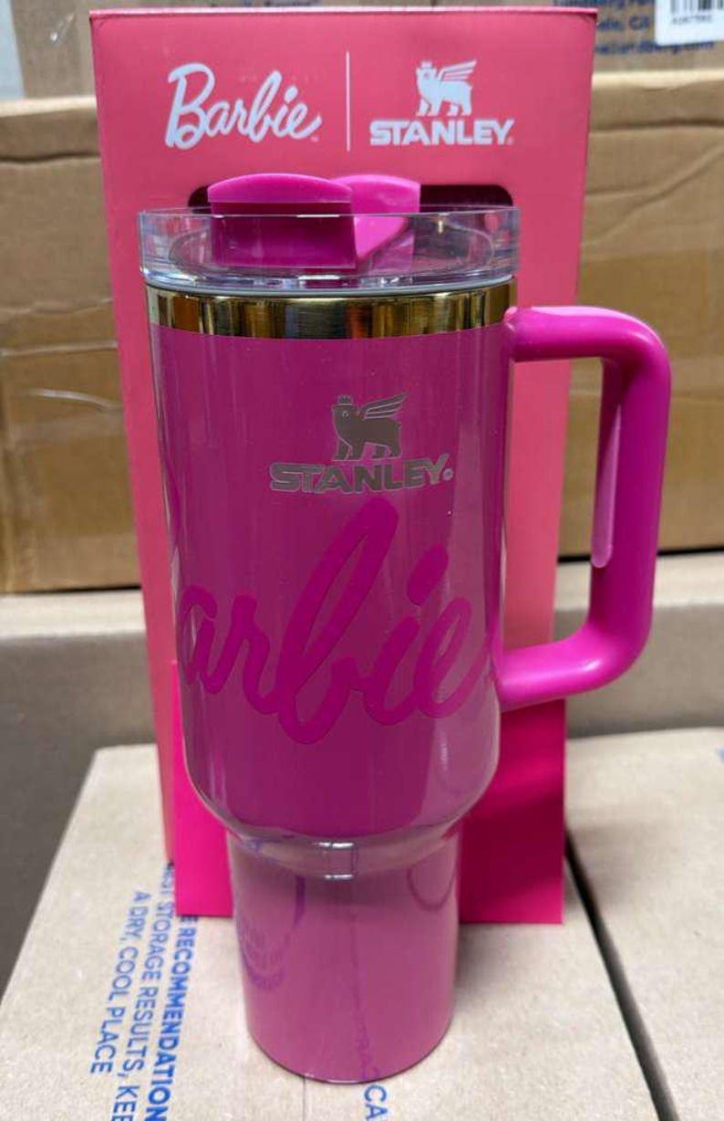 May include: A pink Stanley travel mug with the Barbie logo. The mug has a handle, a clear lid, and gold trim. The Stanley logo is also visible. The mug is in its original pink box.