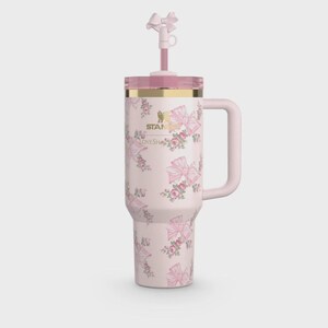 Puede incluir: Un vaso t&eacute;rmico rosa con asa, tapa y pajita, adornado con un estampado floral y de lazo. El vaso tiene un borde dorado y el texto "STANLEY LOVESHACK". La pajita est&aacute; rematada con un lazo.