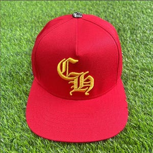 May include: A bright red baseball cap featuring a prominent gold embroidered gothic-style CH monogram on the front panel. The cap includes a small, circular metallic button detail at the very top center of the crown, providing a distinct accent.