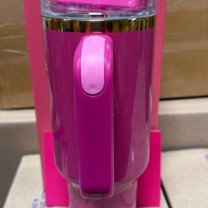 May include: A pink insulated tumbler with a handle, featuring the "Barbie" and "Stanley" logos. The tumbler has a clear lid and a gold-colored band near the top. The handle is a lighter shade of pink. The tumbler is set against a pink box.