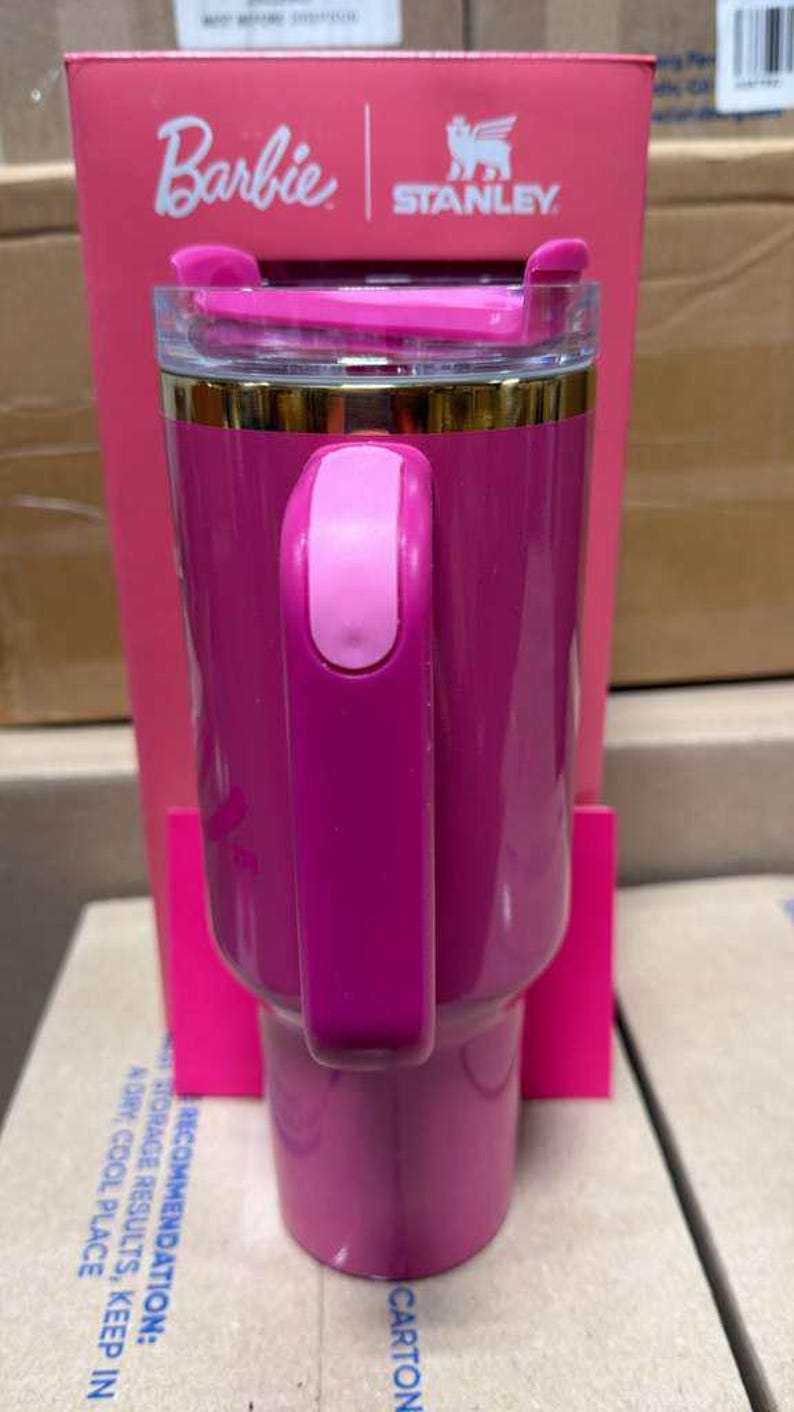 May include: A pink insulated tumbler with a handle, featuring the "Barbie" and "Stanley" logos. The tumbler has a clear lid and a gold-colored band near the top. The handle is a lighter shade of pink. The tumbler is set against a pink box.