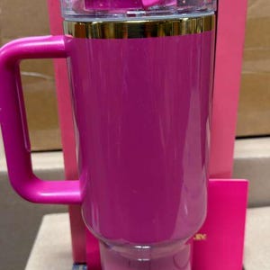 May include: A pink Stanley travel mug with a handle, gold trim, and a clear lid. The mug is part of a Barbie collaboration, with the Barbie and Stanley logos visible on the packaging.