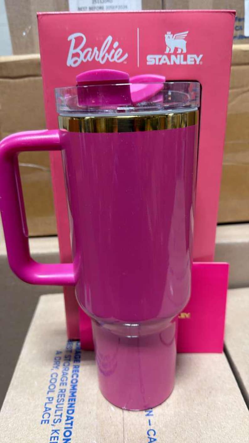 May include: A pink Stanley travel mug with a handle, gold trim, and a clear lid. The mug is part of a Barbie collaboration, with the Barbie and Stanley logos visible on the packaging.