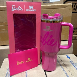 May include: A pink Stanley travel mug with the Barbie logo and a matching pink box. The mug has a handle and a clear lid. The box features the Barbie and Stanley logos. A pink card with the Barbie and Stanley logos is also visible.