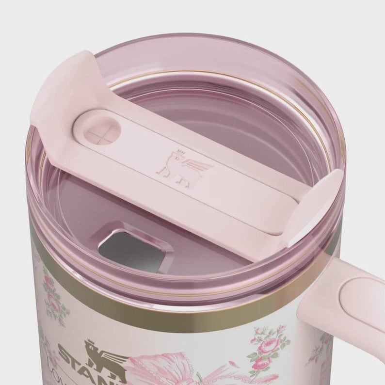 Puede incluir: Un termo de viaje Stanley rosa y blanco con un dise&ntilde;o floral. El termo tiene una tapa rosa y un cierre deslizante rosa con un logotipo de cerdo. El termo tiene un asa y una banda dorada en el medio.
