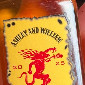 May include: A bottle of Wedding Whiskey with a red cap and a yellow label. The label features the names "Ashley and William", a red graphic, and the text "Wedding Whiskey". The bottle contains a light brown liquid.