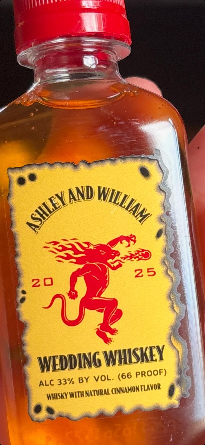 May include: A bottle of Wedding Whiskey with a red cap and a yellow label. The label features the names "Ashley and William", a red graphic, and the text "Wedding Whiskey". The bottle contains a light brown liquid.