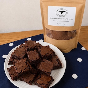 May include: A white plate piled with square chocolate fudge sourdough brownies next to a brown paper pouch labeled Austin Born & Bread Chocolate Fudge Sourdough Brownies, Naturally inspired, no maintenance required, Net Wt. 24 oz (675 g).