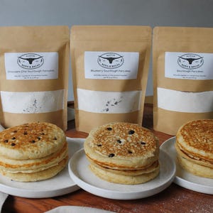 May include: Three brown paper pouches labeled Austin Born & Bread containing pancake mix, positioned behind three white plates each holding a stack of cooked pancakes. The flavors include chocolate chip, blueberry, and plain sourdough pancakes.