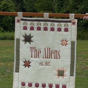 May include: A rectangular quilted wall hanging featuring a cream-coloured centre with the text The Allens and est. 2027 in brown. The border displays a pattern of maroon tulips, olive green star motifs, and dark brown accents on a cream background.