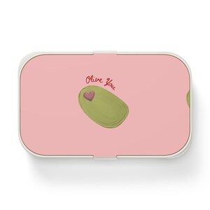 Puede incluir: Una fiambrera rectangular rosa con un borde blanco. La fiambrera presenta una aceituna verde con un corazón rosa y las palabras "Olive You" en escritura roja.