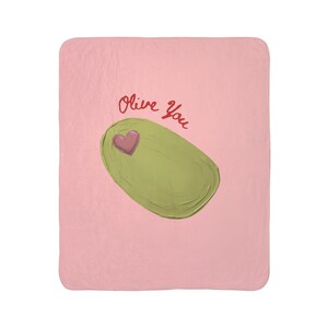 Puede incluir: Una manta rosa con una ilustración de aceituna verde y un corazón rosa. Las palabras "Olive You" están escritas en guion rojo sobre la aceituna. La manta es un artículo suave y acogedor.