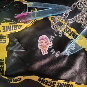 Può includere: Una superficie in pelle nera con nastro giallo della scena del crimine, una catena d'argento e un adesivo di un personaggio che tiene un'arma. L'immagine presenta anche un paio di forbici con una lama trasparente e un piccolo coltello.