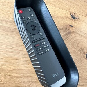 Peut inclure: Une télécommande de télévision LG noire est placée dans un support noir, nervuré, imprimé en 3D. La télécommande comporte divers boutons, dont l'alimentation, le volume et les raccourcis des services de streaming. Le support est sur une surface en bois.