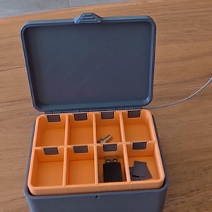 Peut inclure: Une boîte de rangement rectangulaire grise avec un couvercle à charnière ouvert et un intérieur orange vif divisé en huit compartiments individuels. La boîte contient une petite clé USB noire, une vis métallique et deux petits composants carrés gris.