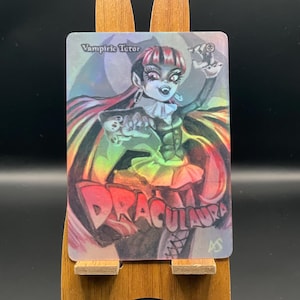 May include: A holographic trading card featuring the character Draculaura from Monster High. The card has the text "Vampiric Tutor" at the top and "Draculaura" in large letters. The artwork depicts Draculaura in a pose with a bat.