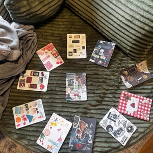 May include: A collection of decorative rectangular stickers with various designs, including cherries, seashells, and abstract art. The stickers are arranged on a textured green surface, with a pillow and blanket in the background.