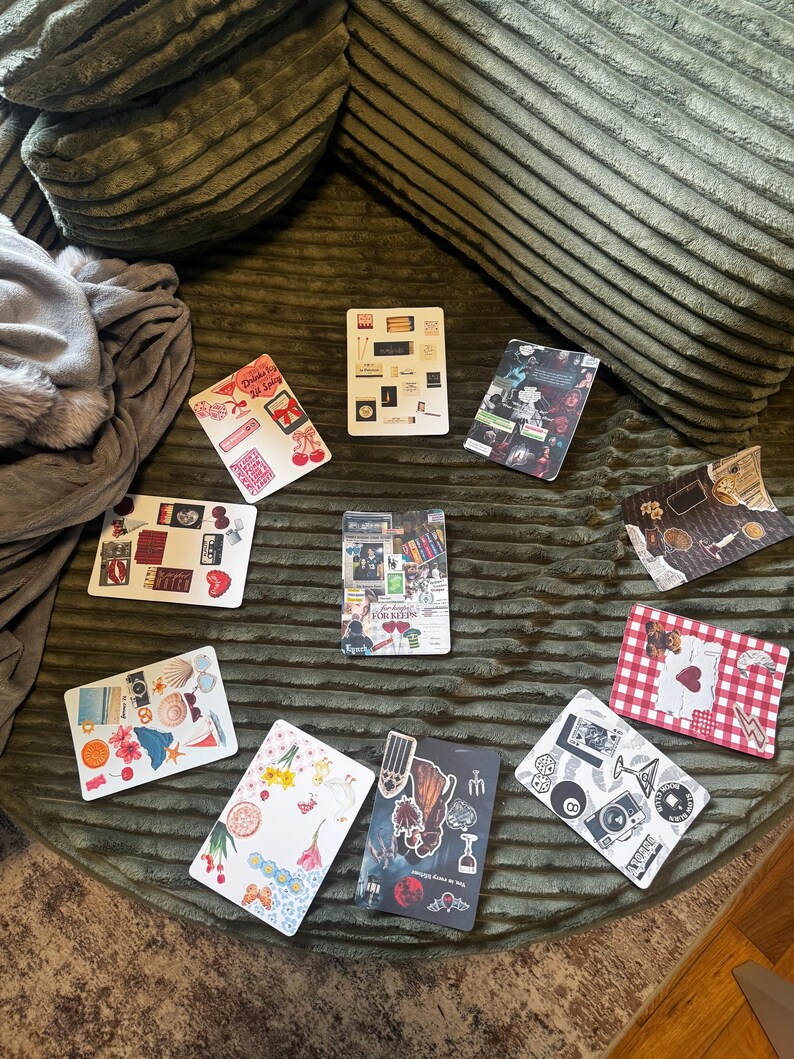 May include: A collection of decorative rectangular stickers with various designs, including cherries, seashells, and abstract art. The stickers are arranged on a textured green surface, with a pillow and blanket in the background.