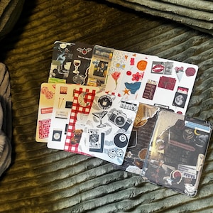 May include: A collection of decorative notebooks with various designs. The notebooks feature images of cocktails, dice, flowers, and vintage-style illustrations. The color palette includes red, white, black, and brown. The notebooks are arranged on a green textured surface.