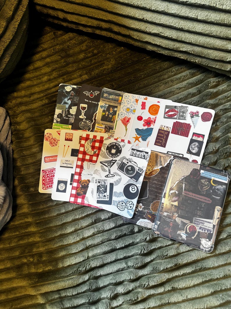 May include: A collection of decorative notebooks with various designs. The notebooks feature images of cocktails, dice, flowers, and vintage-style illustrations. The color palette includes red, white, black, and brown. The notebooks are arranged on a green textured surface.