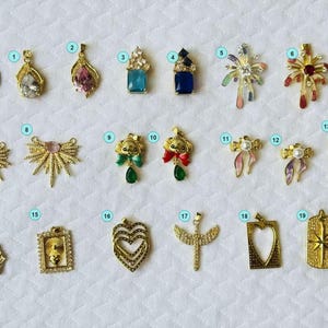 May include: A collection of nineteen gold-toned metal pendants numbered one through nineteen, featuring various shapes including hearts, stars, bows, and geometric designs with colorful accents, displayed alongside a US quarter coin for scale.
