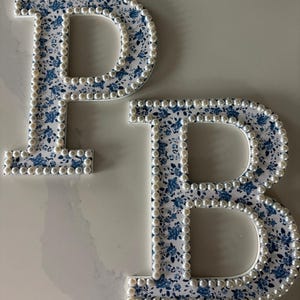 May include: Two decorative letters, P and B, featuring a white surface with a blue floral pattern. Each letter is outlined with a border of small, round, white faux pearls that follow the contours of the characters, creating a textured edge.
