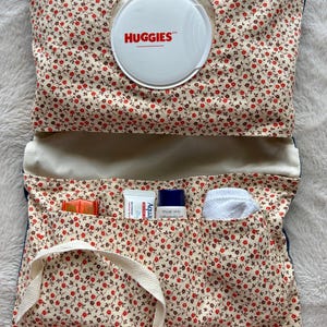 Może przedstawiać: Etui na chusteczki nawilżane w kwiatowy wzór z białą pokrywką z napisem "HUGGIES". Etui jest otwarte, odsłaniając kieszenie z małymi tubkami, małą białą szmatką i małym pomarańczowym przedmiotem. Widoczny jest biały pasek.