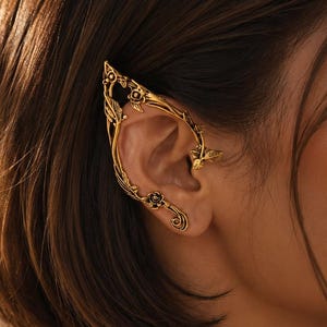 May include: A gold-toned ear cuff featuring an intricate design of sculpted leaves and small flowers that wraps around the outer edge of the ear. The metal piece follows the natural curve of the ear cartilage, creating a detailed botanical appearance.
